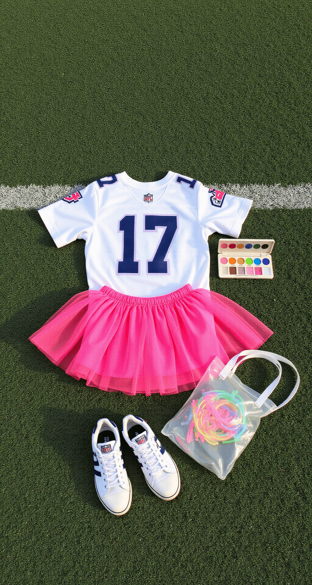 Flat lay arrangement on football field turf featuring a white jersey, neon pink tutu, light-up shoelaces, face paint palette, and a clear stadium bag overflowing with glow sticks, captured in natural midday lighting from an overhead angle.