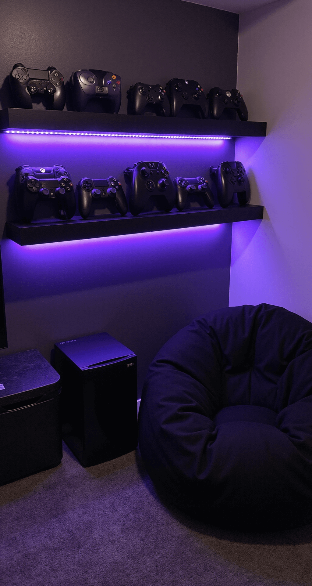 A cozy black gaming corner featuring a plush oversized bean bag on a dark gray rug, a floating shelf with neatly arranged black game controllers highlighted by purple LED strips, and a matte black mini-fridge on a side table, illuminated in the soft light of early evening.
