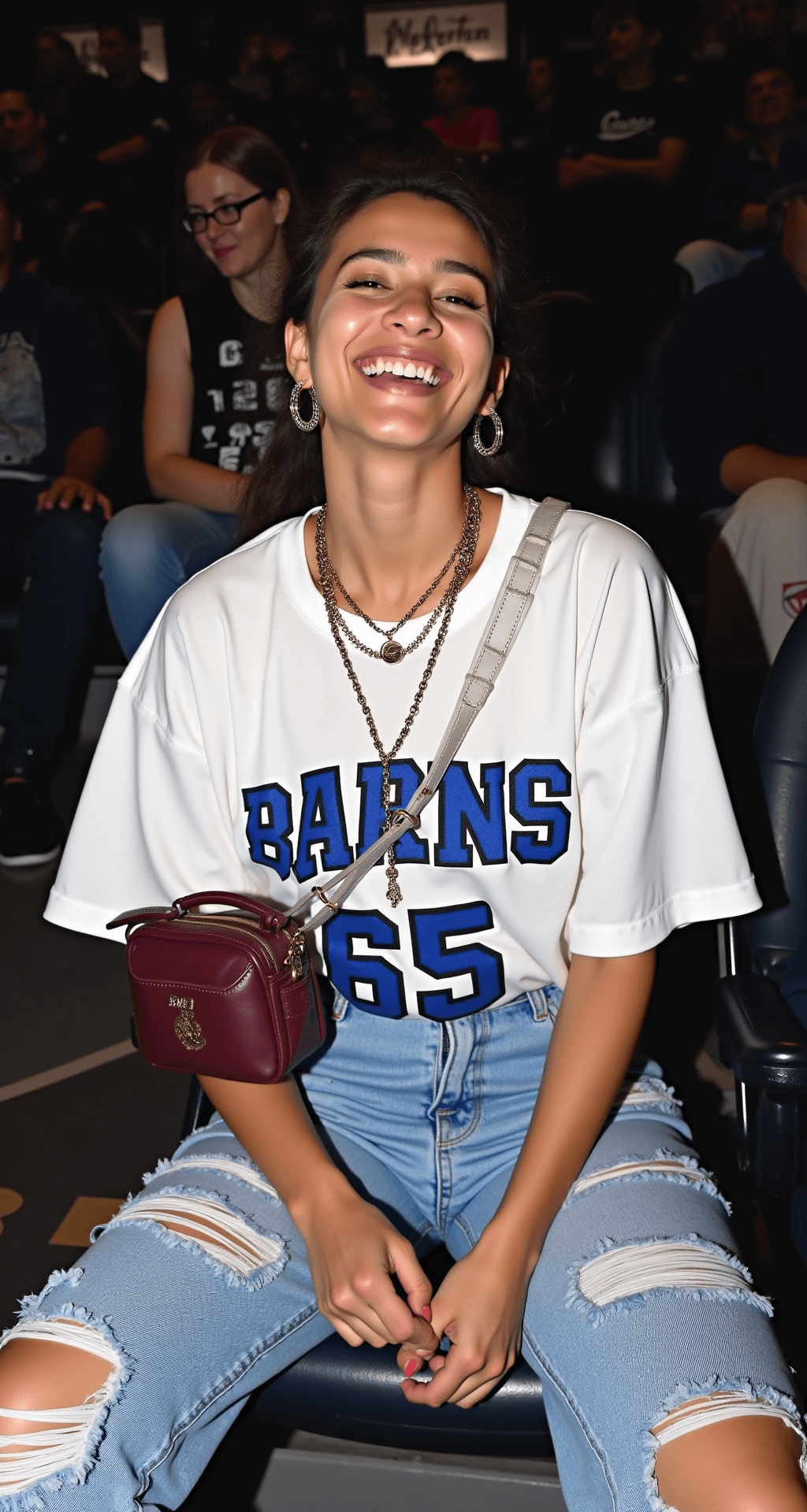 Game Day Glam: Your Ultimate Guide to Basketball Game Outfits (That Actually Work) A model laughing while seated in court-side arena seats, dressed in distressed boyfriend jeans, a cropped white team jersey, and classic Jordan 1s in university blue. She accessorizes with layered gold necklaces, small hoop earrings, and a compact burgundy leather crossbody bag. The shot, taken from slightly above, captures the ambient lighting of stadium lights and emphasizes her natural, glowing makeup and casual style.