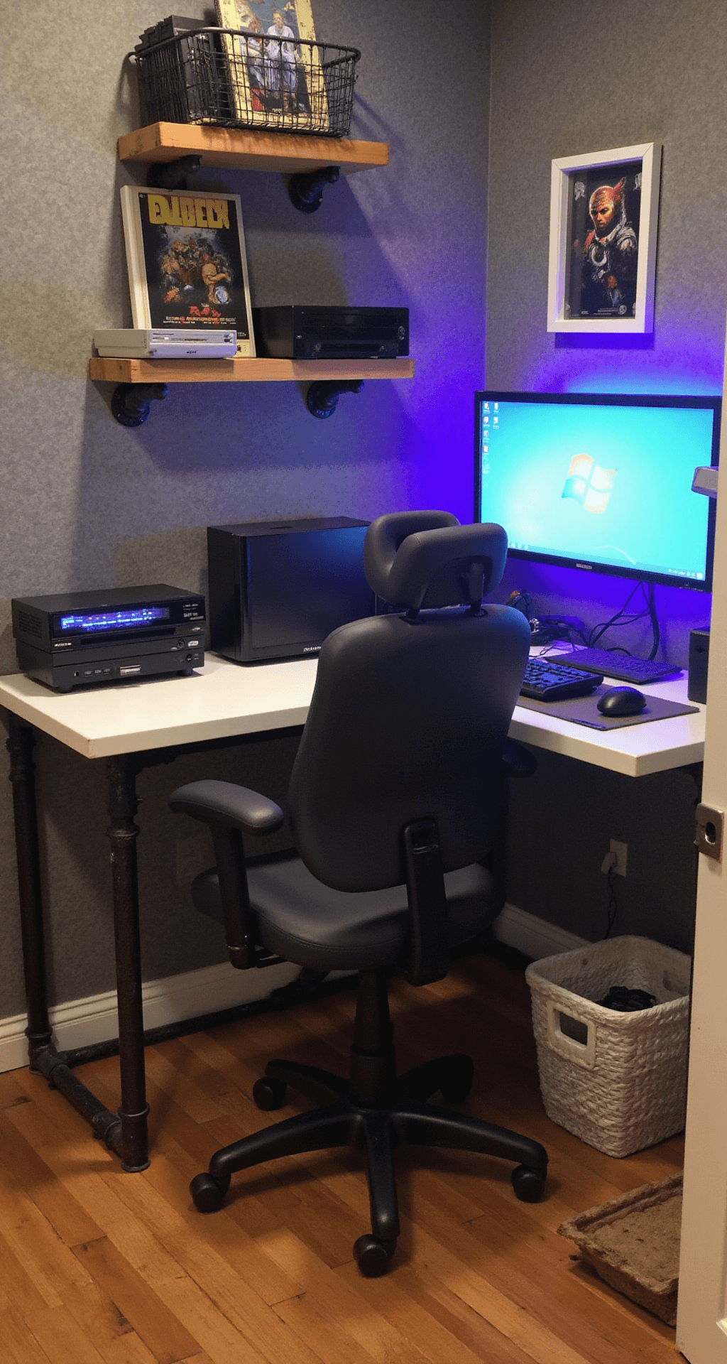 A cozy 12x12ft gaming corner featuring a DIY floating desk with an IKEA countertop and steel legs, a refurbished gaming chair with new upholstery, and LED strips providing ambient lighting. A second-hand monitor is mounted on a cost-effective arm, while wire basket cable management keeps the area tidy. Vintage gaming posters in simple frames adorn the walls, and industrial pipe shelving showcases gaming consoles.