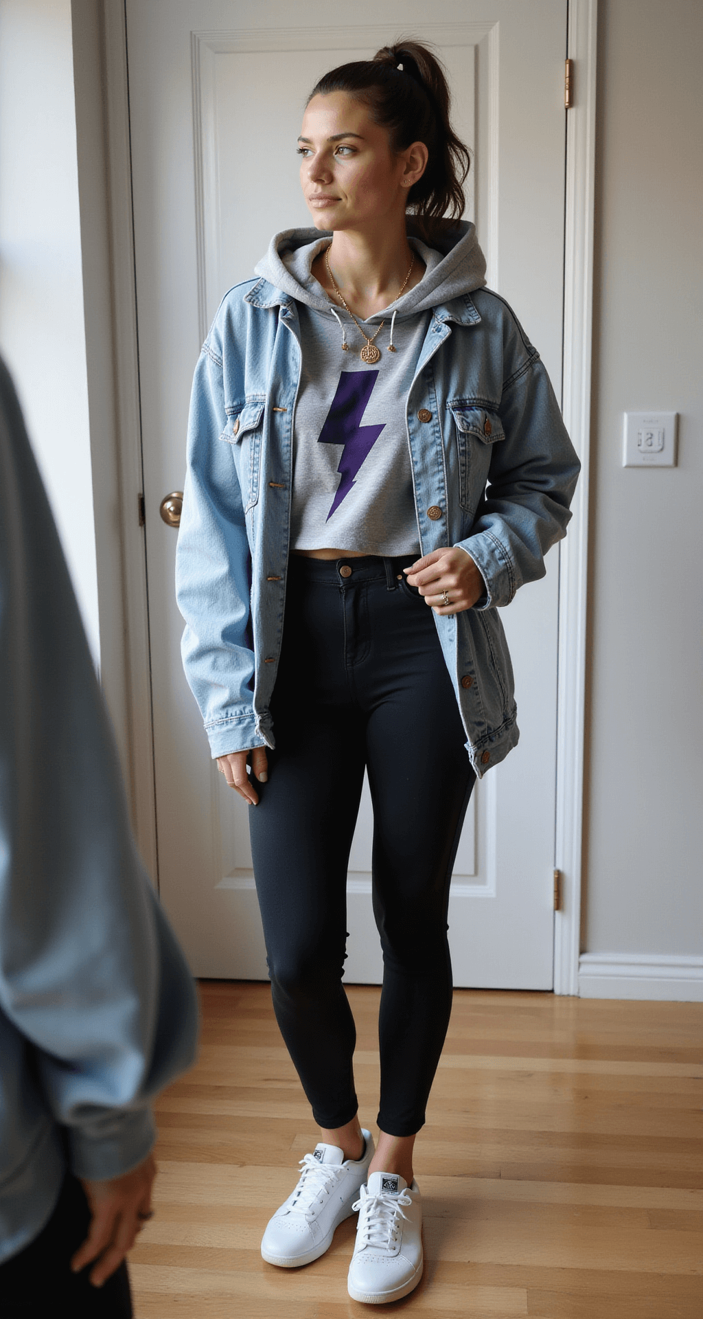 What to Wear to a Basketball Game: The Ultimate Style A modern apartment interior featuring a pre-game scene with a person styling layers at a full-length mirror, wearing black high-rise leggings, a cropped heather gray team hoodie, and an oversized light wash denim jacket. Accessories include a lightning bolt pendant necklace and small gold hoop earrings. White Reebok Classics are positioned by the door, illuminated by soft window light for an intimate documentary feel.