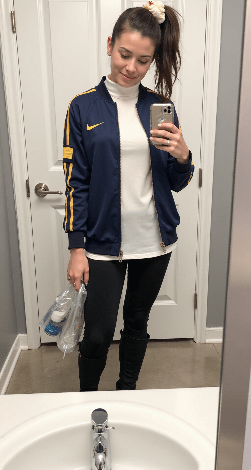 The Perfect Soccer Game Outfit: Look Amazing While Cheering on Your Team! A bathroom mirror selfie in a modern stadium, featuring a person in a fitted jersey over a white turtleneck, black leggings, and ankle boots. The hair is styled in a sleek high ponytail with a team scrunchie. The clear bag reflects organized game day essentials, with bright, even lighting highlighting the outfit's proportions.
