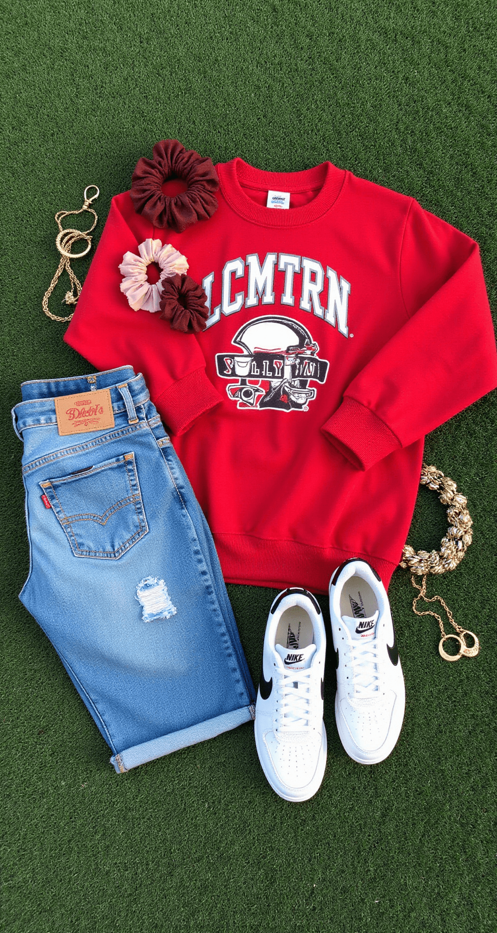 The Ultimate Guide to High School Football Game Fashion: Look Cute & Stay Comfy Flat lay of a game day outfit on green turf featuring an oversized team sweatshirt, distressed Levi's mom jeans, white Nike Air Force 1s, minimal gold jewelry, and team-colored scrunchies, captured in natural daylight.