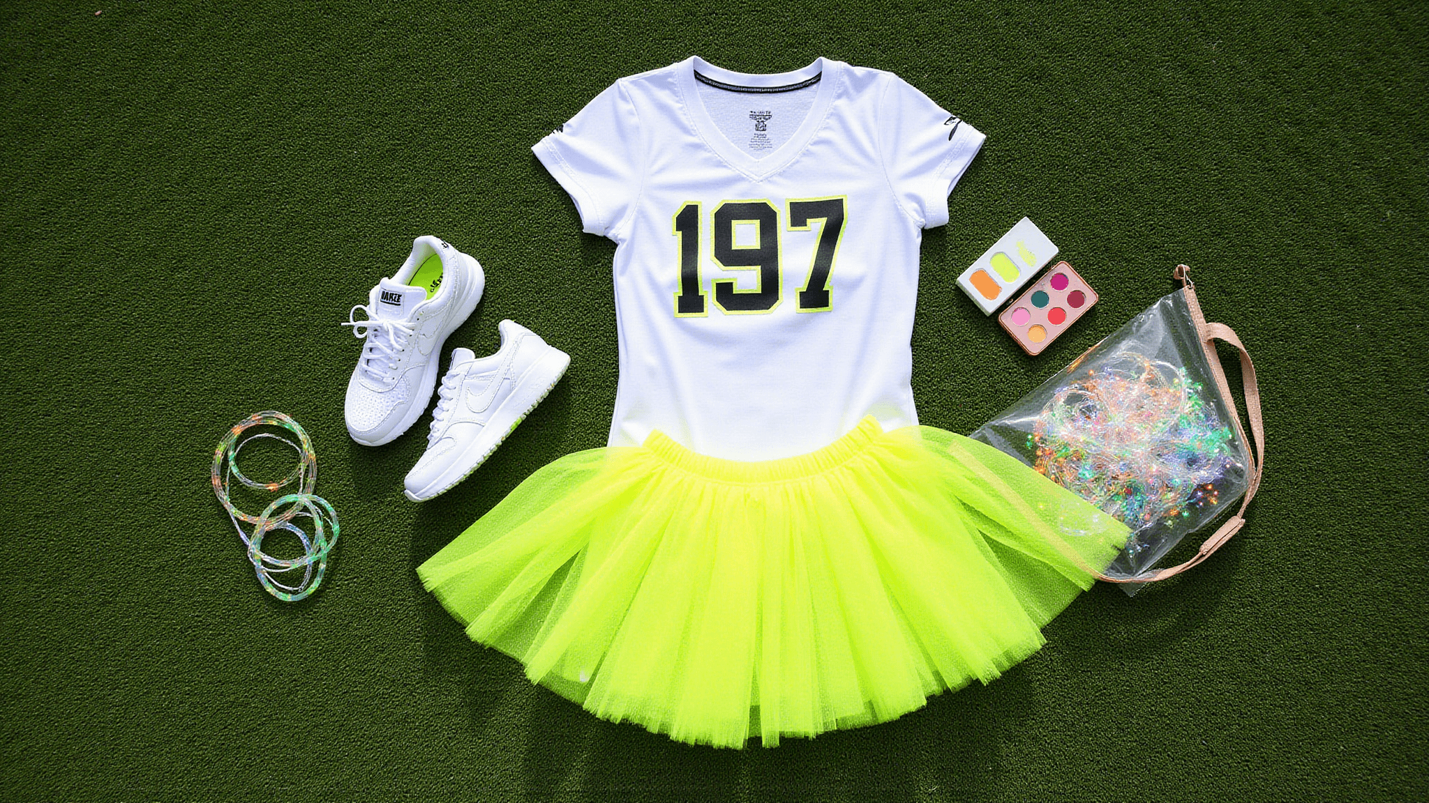 Flat lay of game day outfit on green football turf, featuring a white jersey, neon yellow tutu, light-up Nike sneakers, glow stick bracelets, a face paint palette, and a clear stadium bag with fairy lights, all captured in bright natural light.