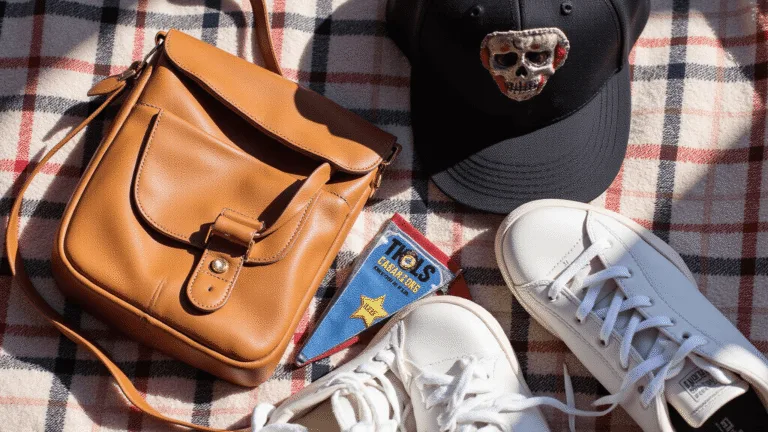 A flat lay of vintage sports fan accessories on a plaid blanket, including a leather crossbody bag, a distressed baseball cap, team pennants, and white sneakers, illuminated by warm afternoon sunlight.