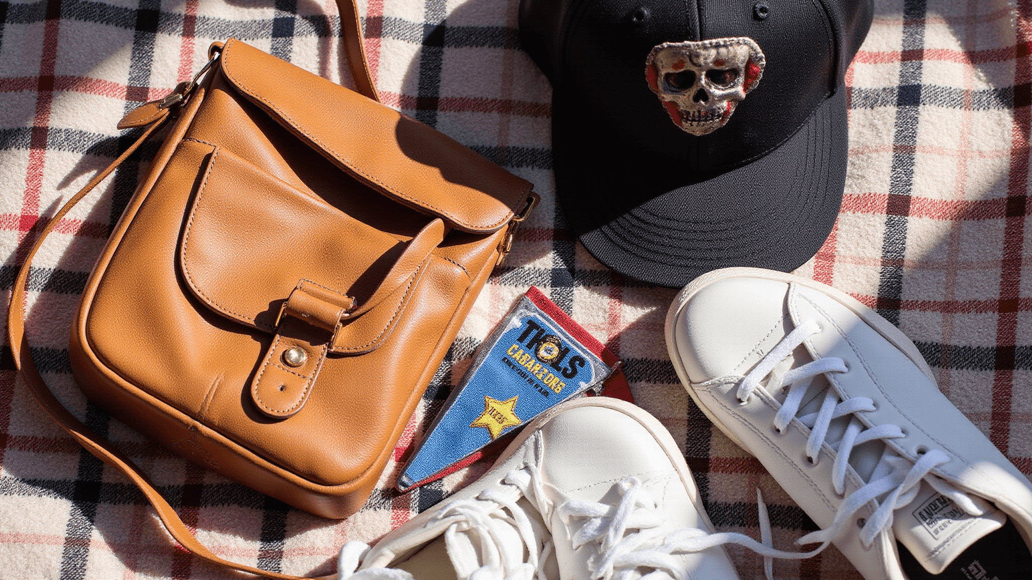 A flat lay of vintage sports fan accessories on a plaid blanket, including a leather crossbody bag, a distressed baseball cap, team pennants, and white sneakers, illuminated by warm afternoon sunlight.